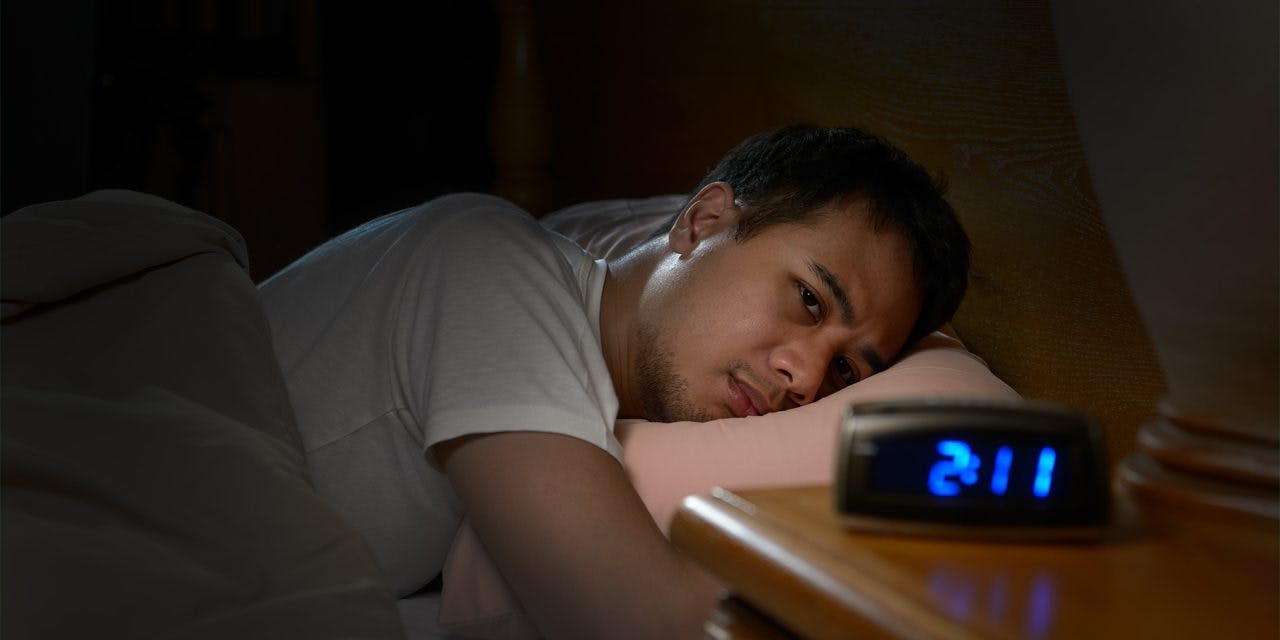 hombre tumbado en su cama con los ojos abiertos frente a un reloj digital