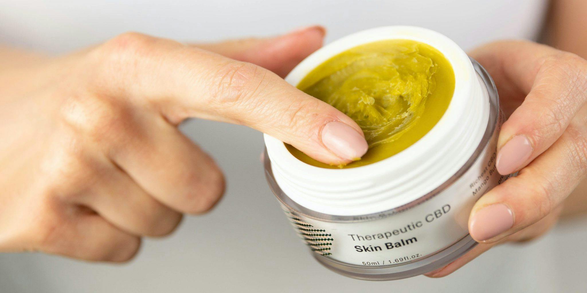 closeup woman's pair of hands holding a cannabis topical cream, one finger rubbing the cream