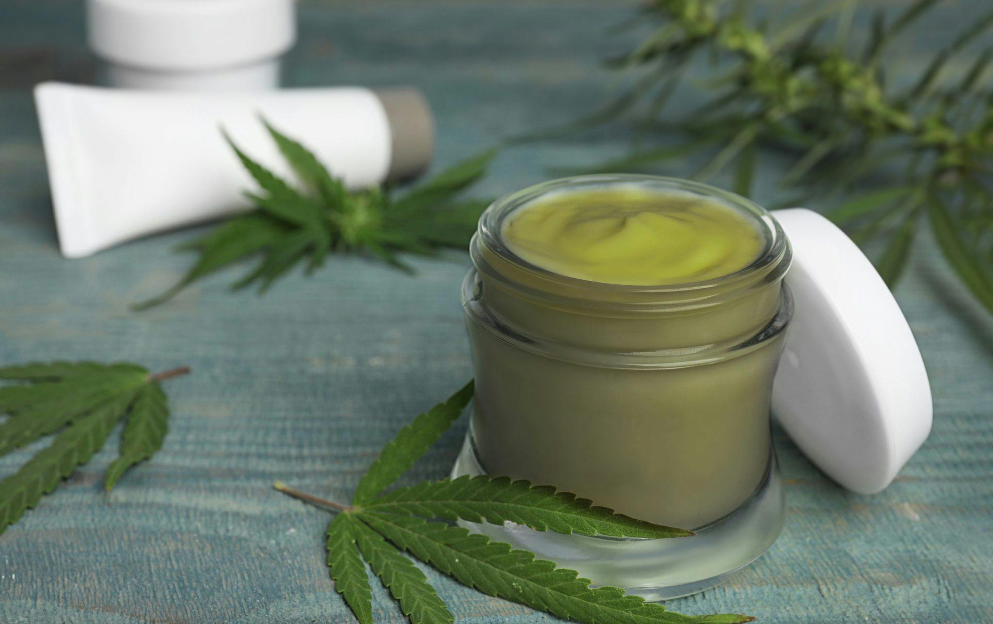 closeup cannabis topical on a small jar with cannabis leaves and cream tube in the background
