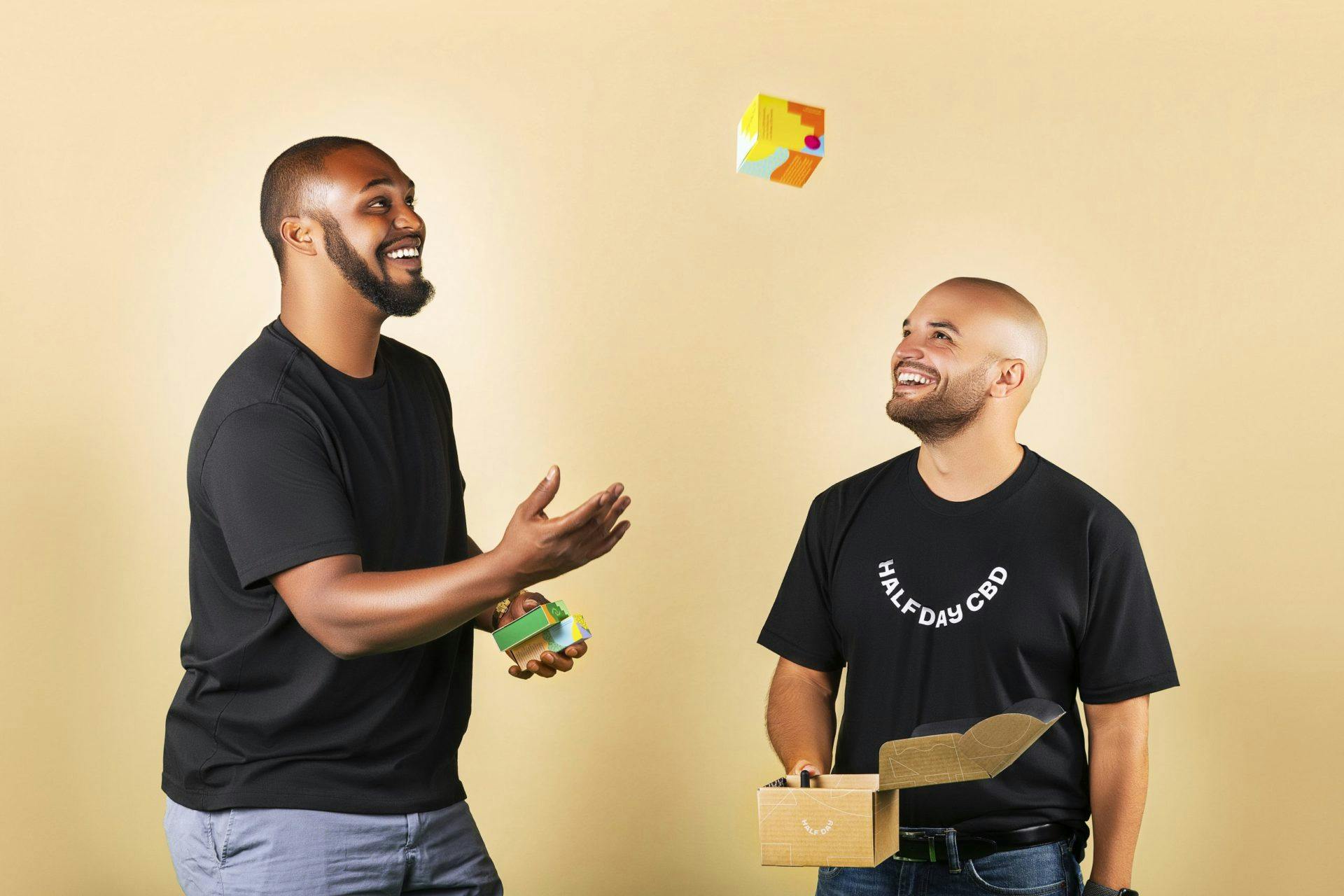 two bald men wearing black shirts, tall man tossing small box while the other holds empty opened box