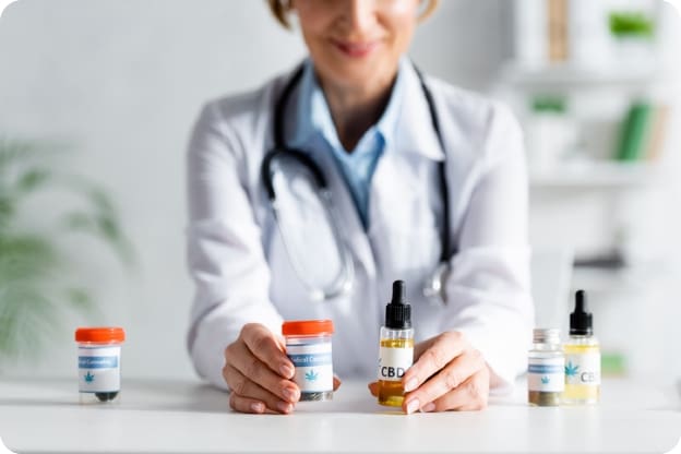 Female doctor sits across table holding CBD in pill bottle in one hand and CBD oil in a dropper bottle in another